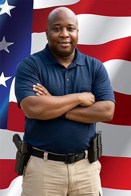 Founder and Chief Training Instructor Theodore Hampton II standing in front of an American Flag with arms crossed wearing a holster handgun and magazines n a belt.